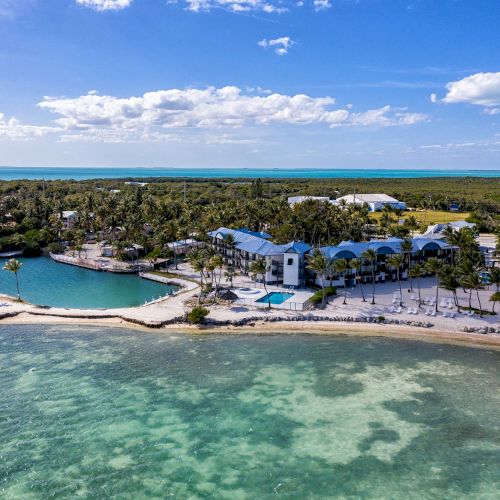 Aerial view of a coastal resort with turquoise waters, a pool, and palm trees, under a blue sky with scattered clouds.