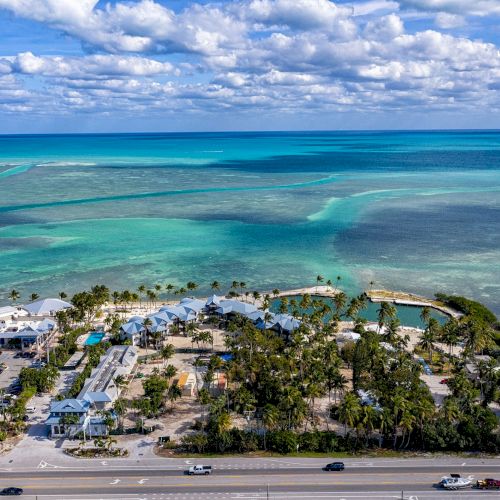 An aerial view of a coastal area with buildings, palm trees, a road, and the turquoise ocean in the background under scattered clouds.