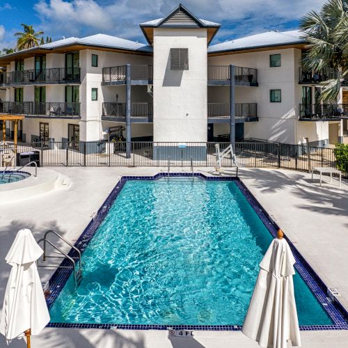 An outdoor swimming pool with lounge chairs and umbrellas, adjacent to a building surrounded by palm trees, under a partly cloudy sky.