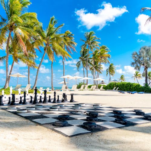 A large outdoor chessboard on a sandy beach, surrounded by palm trees, blue sky, and ocean view. Lounge chairs are in the background.