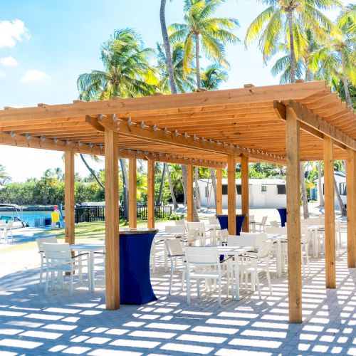 The image shows a wooden pergola with white chairs and tables beneath, situated outdoors near a pool and palm trees, on a sunny day.