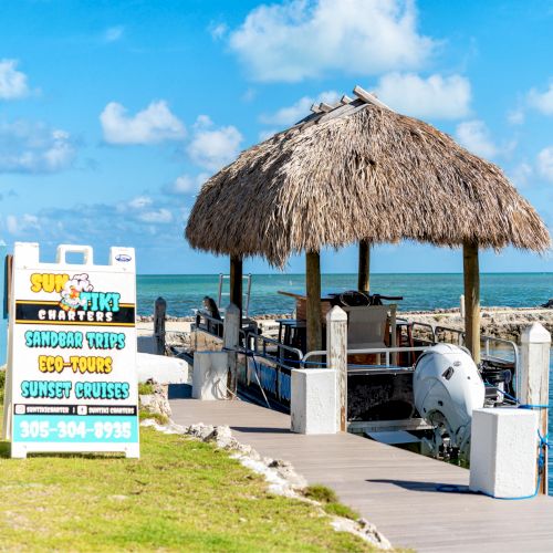 A tropical dock with a thatched hut, a boat, and a sign offering sandbar trips, eco-tours, and sunset cruises by Sun & Fun Charters.