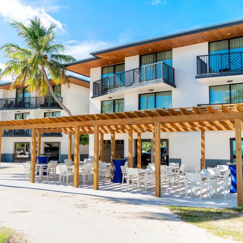 A modern building with balconies, a wooden pergola over outdoor seating, and palm trees under a blue sky.