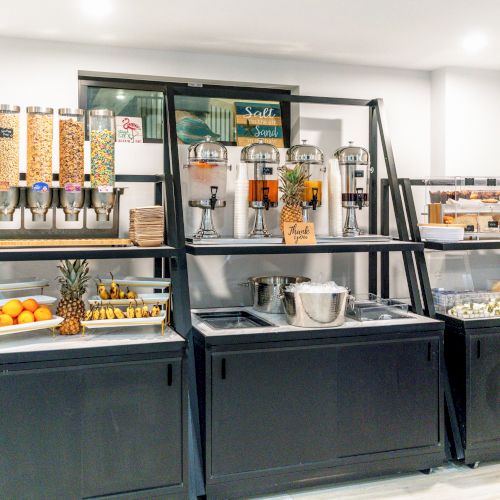 A breakfast buffet with cereal dispensers, a juice machine, fresh fruit, and pastries, neatly arranged on black stands.