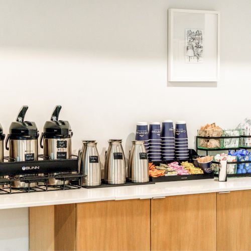 A coffee station with dispensers, cups, snacks, and creamers on a countertop under a simple framed artwork.