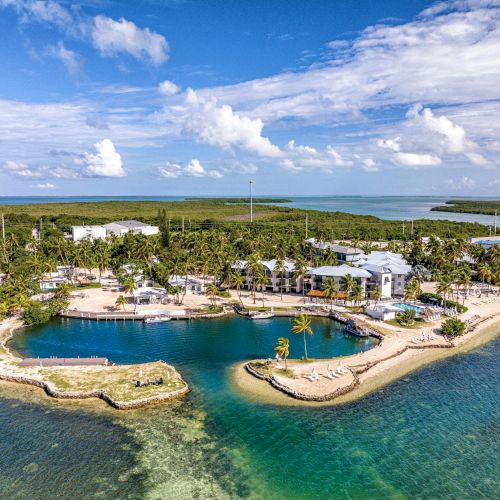 Aerial view of a coastal resort area with clear blue water, sandy beaches, and lush greenery under a partly cloudy sky.