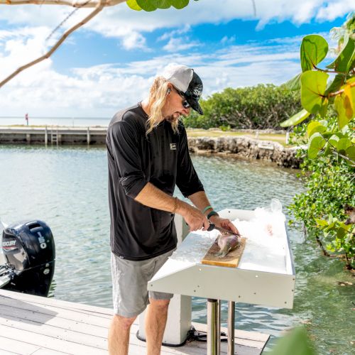 A person is cleaning a fish on a table by the water, surrounded by greenery, with a dock and a motorboat nearby.