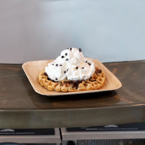 A waffle topped with whipped cream and chocolate chips on a square plate, placed on a curved metallic surface.