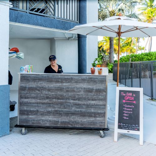 Outdoor bar setup with a wooden counter, an overhead umbrella, a staff member, and a sign announcing the bar is open.