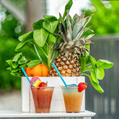 Two tropical drinks with straws and fruit garnishes sit in front of a box with a pineapple, leaves, and oranges, outdoors.