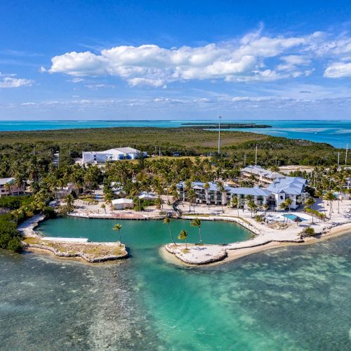 Aerial view of a coastal resort with clear blue water, sandy beaches, and surrounding greenery under a partly cloudy sky.