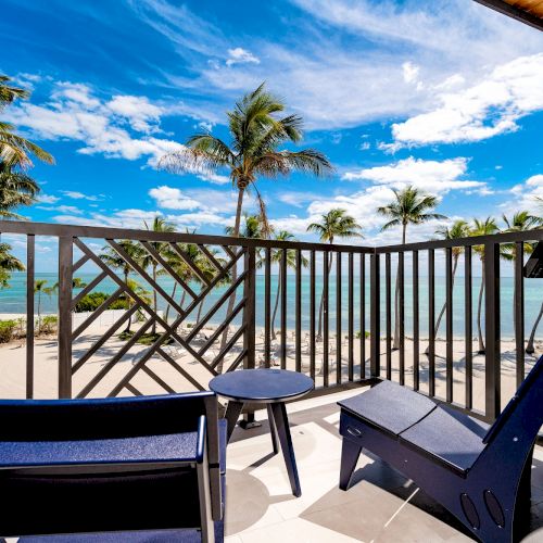 A balcony overlooking a tropical beach scene with palm trees, blue skies, and ocean. Two modern chairs and a small table are present.