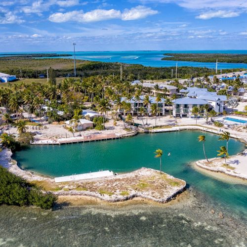 Aerial view of a tropical island with clear water, sandy beaches, lush greenery, and buildings, connected by a narrow road or bridge.