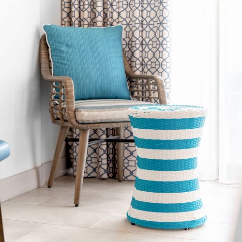 A wicker chair with a blue cushion next to a blue and white striped wicker stool, with patterned curtains in the background.