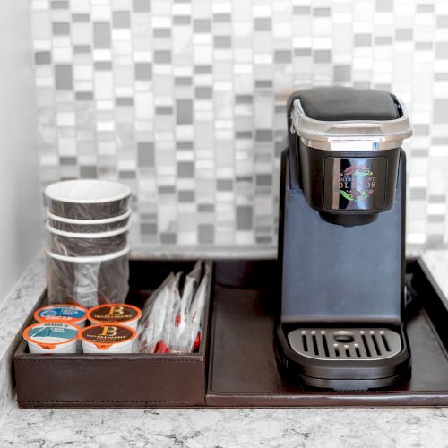 A small coffee maker on a tray with disposable cups, coffee pods, and stirrers, set on a marble countertop against a tiled wall.