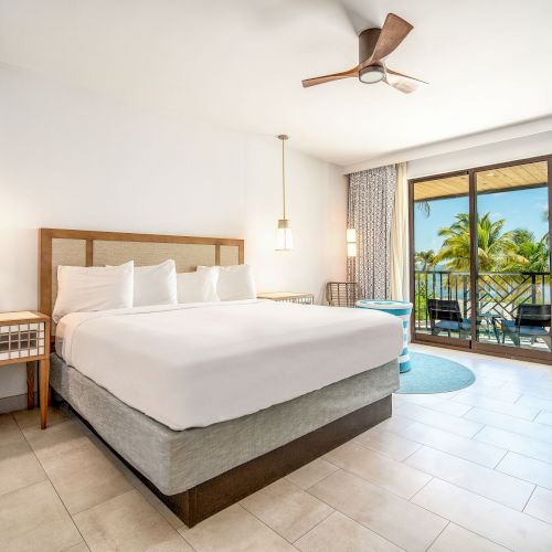 A modern hotel room features a king-sized bed, ceiling fan, lamps, sofa, and a balcony with a view of palm trees and the ocean.