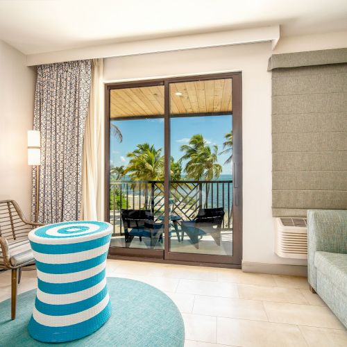 A modern hotel room with a striped stool, chair, and sofa, overlooking a balcony with palm trees through sliding glass doors.