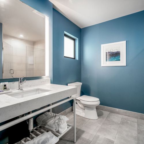 A modern bathroom with blue walls, a white sink, mirror, toilet, and shower. A towel shelf and wall art complete the decor.