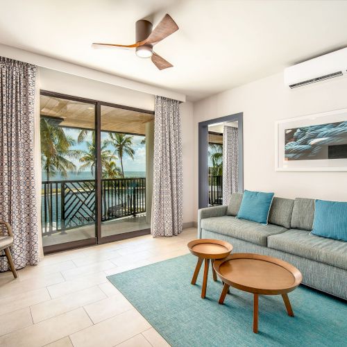 A modern living room with a couch, coffee tables, ceiling fan, and large window view of palm trees and ocean, creating a tropical ambiance.