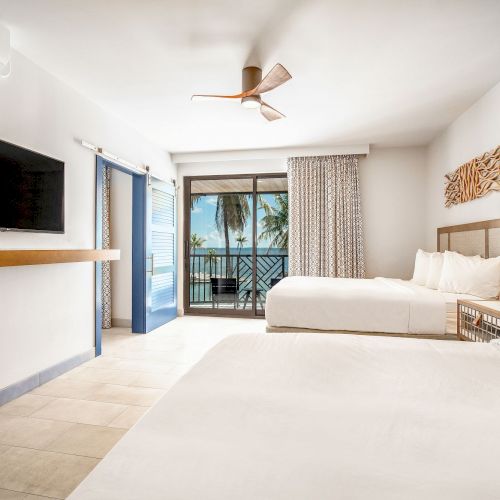 A hotel room with two beds, a TV, ceiling fan, and a balcony with ocean views through sliding doors.