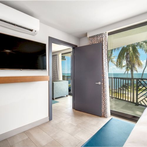 A bright room with a wall-mounted TV, open door to a balcony, ocean view with palm trees, tiled floor, air conditioner on the wall.