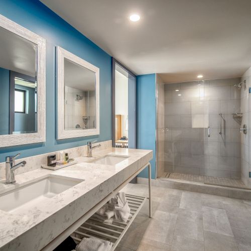 A modern bathroom with blue walls, double sinks, a large mirror, and a spacious shower area with glass doors.