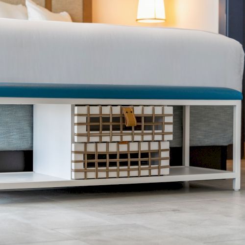 A modern bench with a blue cushion and a decorative basket underneath, located at the foot of a bed in a neatly furnished room.