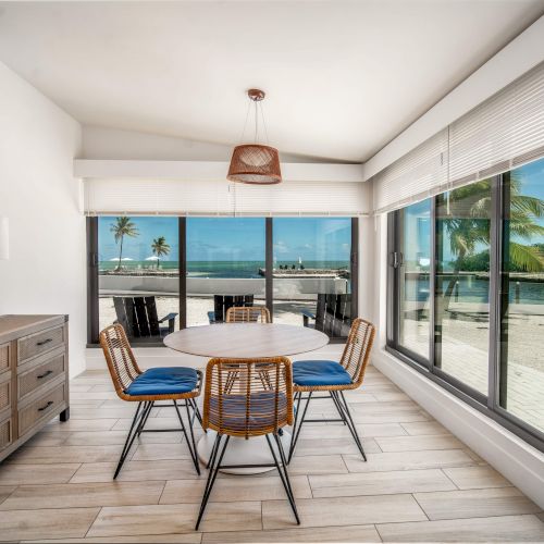 A dining room with a round table, wicker chairs, large windows showing a tropical view with palm trees and water.