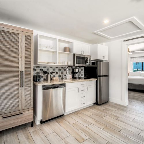 A modern kitchenette with wooden cabinets, a fridge, microwave, and sink, leads to a bedroom with a neatly made bed through a sliding door.