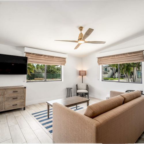 A modern living room with a beige sofa, striped rug, TV, fan, and wall art, featuring large windows and a wooden floor.