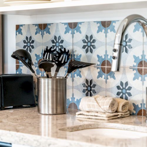 A kitchen counter with a sink, a polished faucet, utensils in a holder, a toaster, towels, and decorative blue patterned tiles adorn the wall.