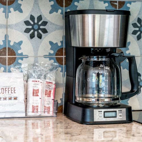 A coffee maker on a counter with a packet of coffee and creamer, set against a patterned tile backdrop.