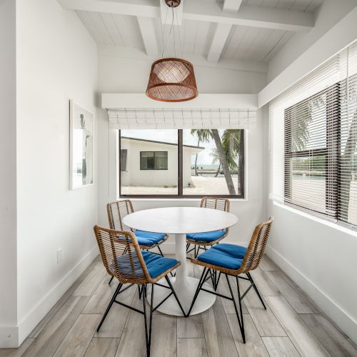 A bright dining area with a round table and four chairs, blue cushions, large windows, and an overhead light fixture, offering a sunny view.