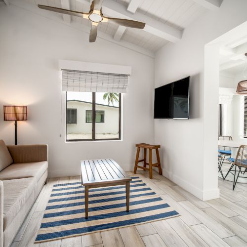 A modern living room with a sofa, striped rug, TV, and ceiling fan, leading to a dining area with a window view.