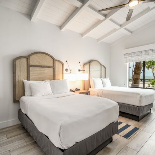 The image shows a bright bedroom with two neatly made beds, a window with a view of trees, and a wooden ceiling fan above.