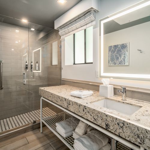 The image shows a modern bathroom with a glass shower, a granite countertop sink, towels, and a backlit mirror on the wall.