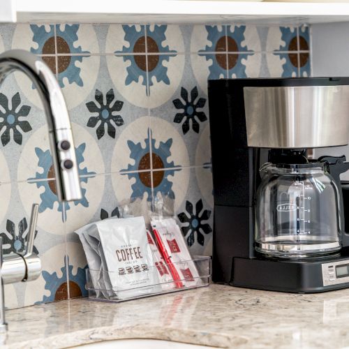 A kitchen counter with a coffee maker, coffee packets, and a stainless steel faucet against a patterned tile backsplash.