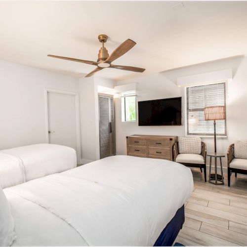 A modern bedroom features two beds, a ceiling fan, a TV, chairs, dresser, and a window, all in a clean, neutral color palette.