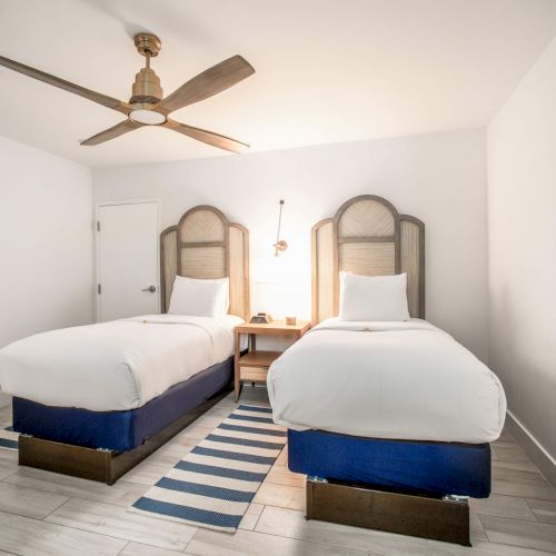 A minimalist bedroom with two single beds, a ceiling fan, and a striped rug on a wooden floor, featuring neutral tones and simple decor.