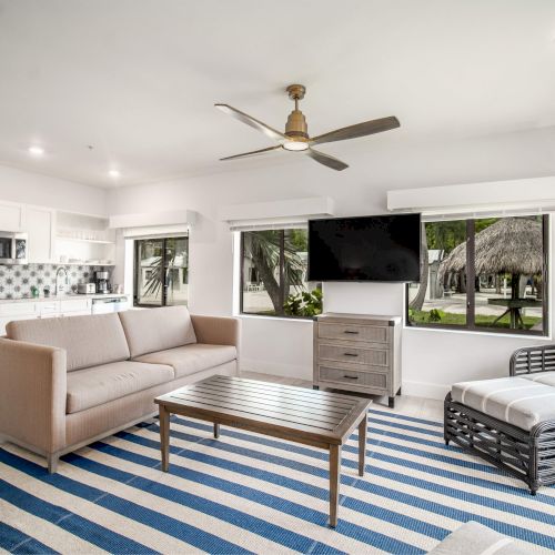 A modern living room with a sofa, striped rug, TV, and windows showing an outdoor view. Kitchen area is visible in the background.