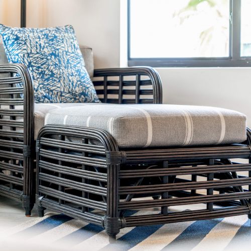 A dark rattan armchair with a gray cushion and a blue patterned pillow, placed on a striped rug by a window.