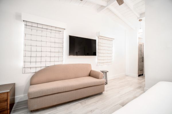 A modern living area with a beige couch, wall-mounted TV, and two windows with white blinds; features wooden flooring and a minimalist design.