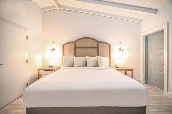 A modern bedroom with a large bed, white linens, two bedside tables, wall-mounted lamps, and a door to the right. The room has a clean, minimalist look.