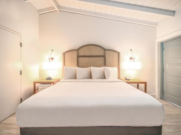 A modern bedroom with a large bed, white linens, two bedside tables, wall-mounted lamps, and a door to the right. The room has a clean, minimalist look.