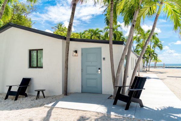 A small white house with a light blue door, surrounded by palm trees, with two black chairs and a small table outside on a sunny day.