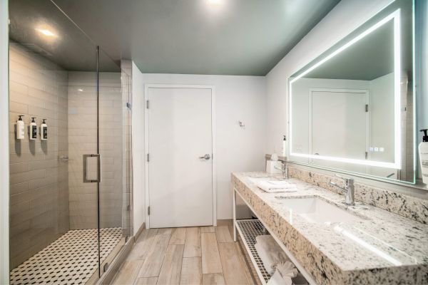 Modern bathroom with a glass-enclosed shower, large vanity, dual sinks, and a backlit mirror on a light wood floor.