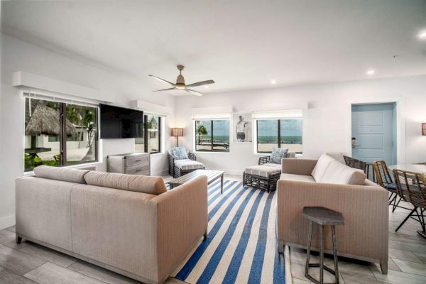 A modern living room with sofas, a blue-striped rug, a ceiling fan, and large windows offering a beach view.