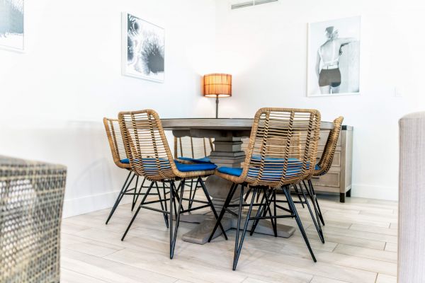 A dining area with a round table, six wicker chairs, a central lamp, and two wall art pieces on a light wooden floor.