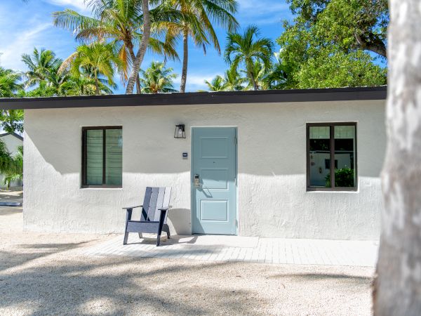 A small white house with a light blue door and two windows, surrounded by palm trees, with a chair placed outside.