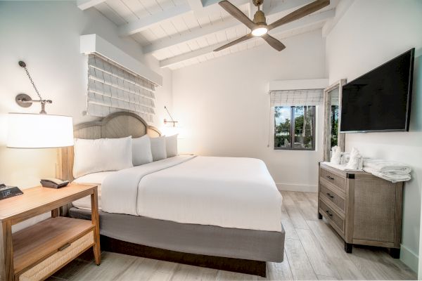 A modern bedroom with a large bed, bedside tables, a ceiling fan, a wall-mounted TV, and a window overlooking greenery.